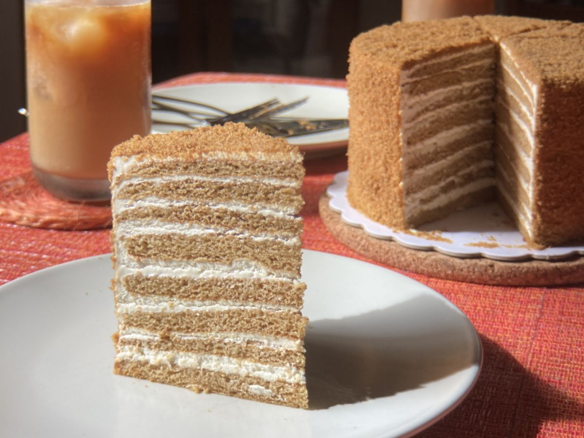 A tall slice of Medovik honey cake with many thin layers, whole cake behind it